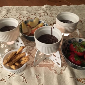 May include: A white ceramic fondue pot with a dark chocolate fondue, surrounded by a white ceramic plate with breadsticks, a white ceramic bowl with apple slices, and a white ceramic bowl with strawberries.