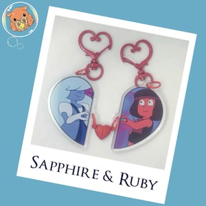 May include: Two heart-shaped acrylic keychains featuring cartoon characters Sapphire and Ruby. Each keychain has a pink heart-shaped clasp and a small red magnetic heart connector. The text Sapphire & Ruby is printed below the keychains.