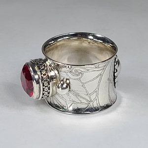 May include: A silver ring featuring a red gemstone and intricate etched designs. The ring is wide and has a textured surface.