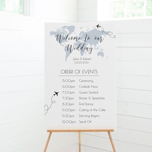 Könnte beinhalten: Ein weißes Schild mit einer Weltkartengrafik und dem Text "Willkommen zu unserer Hochzeit" mit den Namen des Paares und dem Hochzeitsdatum. Das Schild listet die Reihenfolge der Ereignisse für den Hochzeitsempfang auf, einschließlich der Zeremonie, der Cocktailstunde, des Abendessens, der Reden, des Eröffnungstanzes, des Anschneidens der Torte und des Abschlusses.