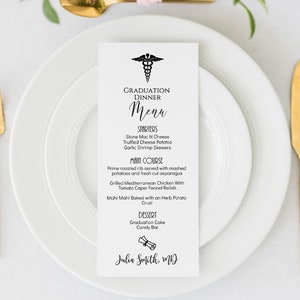 May include: A white graduation dinner menu with a black caduceus symbol at the top. The menu lists starters, main course, and dessert options. The text at the bottom reads "Julia Smith MD".