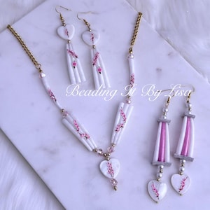 May include: A white necklace and earring set with a floral design. The necklace has a gold chain and a heart-shaped pendant. The earrings have a similar design, with white and pink accents. The text "Beading It By Lisa" is visible.