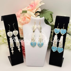 May include: Three pairs of heart-shaped earrings on display. The earrings feature white and light blue hearts with floral designs, gold accents, and dangling elements. The earrings are displayed on black stands and a white mannequin head.