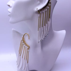 May include: Ear cuffs with a gold-tone finish, featuring dangling white elements, chains, and small pearl accents. The ear cuffs are displayed on a white mannequin head, showcasing their design and details.