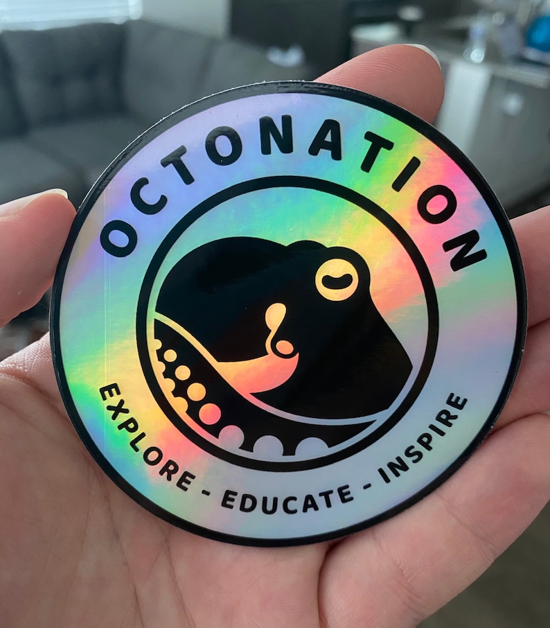 May include: Holographic round sticker with the text "OCTONATION" in black, surrounding a black octopus illustration. The sticker also includes the phrases "EXPLORE - EDUCATE - INSPIRE" in a white font. The sticker has a rainbow iridescent effect.