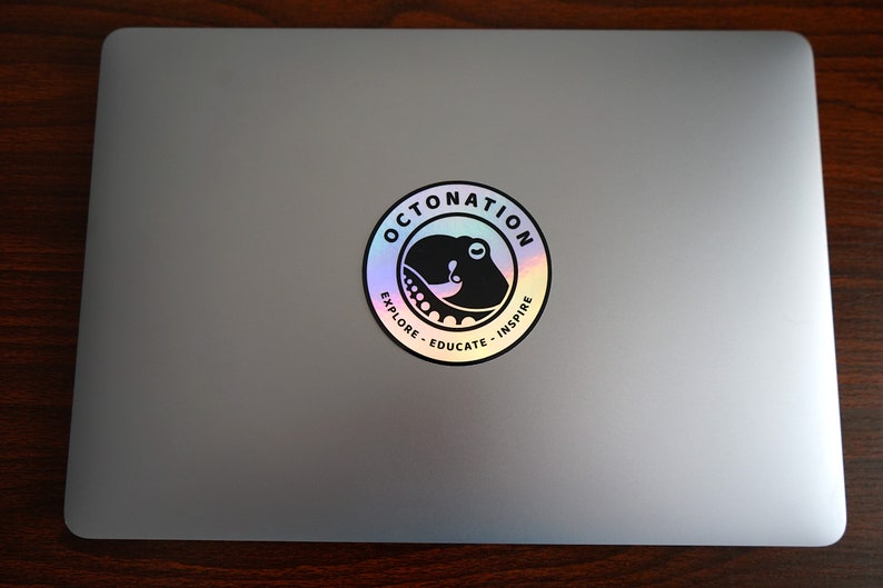 May include: A silver laptop with a circular holographic sticker. The sticker features a black octopus design inside a circle with the words "OCTONATION", "EXPLORE", "EDUCATE", and "INSPIRE" in a white font.