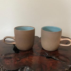 May include: Two ceramic mugs with a matte, earthy brown exterior and a smooth, light blue interior. Each mug features a rounded, looped handle extending from the side. The mugs have a cylindrical shape with a flat base and a simple, clean design.