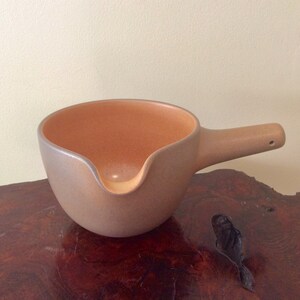 May include: A tan-colored ceramic gravy boat with a handle and a pouring spout. The interior is a lighter shade of tan. The gravy boat is sitting on a dark brown wooden surface.