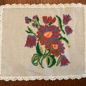 May include: A white crocheted doily with a floral embroidery design. The flowers are purple, red, and green.