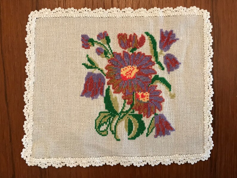 May include: A white crocheted doily with a floral embroidery design. The flowers are purple, red, and green.