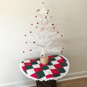 May include: A white feather Christmas tree with red berry ornaments and two small angel ornaments. The tree sits on a wooden base atop a small table covered with a red, green, and white patchwork tree skirt.