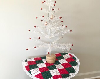 May include: A white feather Christmas tree with red berry ornaments and two small angel ornaments. The tree sits on a wooden base atop a small table covered with a red, green, and white patchwork tree skirt.