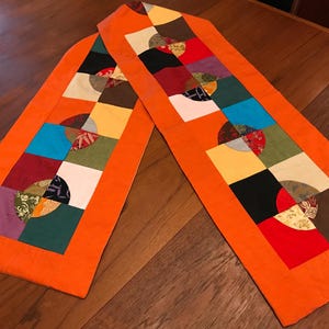 May include: Two orange table runners with a patchwork design. Each runner features a central row of colorful squares, each with a half-circle pattern. The squares are in various colors, including red, blue, green, and black.
