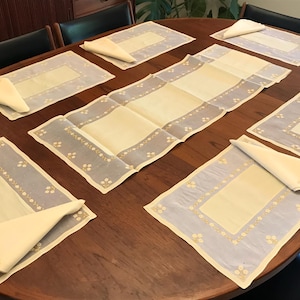 May include: A set of six rectangular placemats and a matching long table runner, all featuring a pale yellow center panel bordered by a light blue frame with a delicate white floral pattern. Each placemat includes a folded cream-colored napkin.