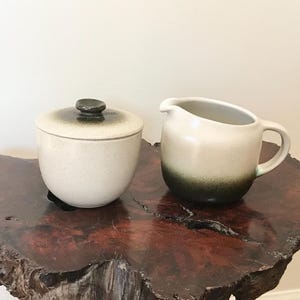 May include: A ceramic sugar bowl with a lid and a small creamer pitcher. Both are off-white with a dark green ombre effect at the base. The sugar bowl has a dark knob on the lid. The items are on a dark wood surface.