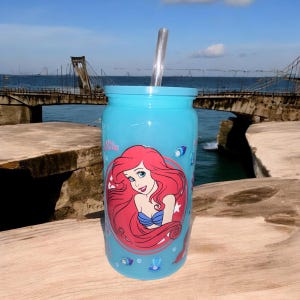 May include: A light blue tumbler with a clear straw. The tumbler features a graphic of Ariel from The Little Mermaid, with her red hair and blue top. The tumbler is decorated with sea-themed illustrations.