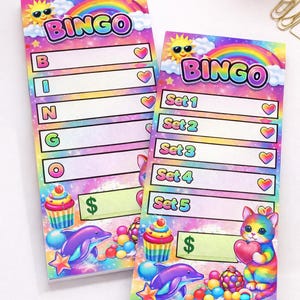 May include: Two colorful Bingo game pads with rainbow, sun, and cloud designs. Each pad features the word "BINGO" at the top, with spaces for numbers and hearts. The bottom of the pads have a cupcake, dolphin, and a cat holding a heart.