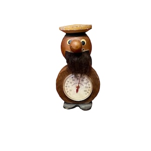 Puede incluir: Un termómetro de madera con una cara de dibujos animados y un bigote. El termómetro tiene una esfera redonda con una esfera negra y blanca y una aguja roja. La esfera indica "Thermometer" y tiene números del 0 al 140.