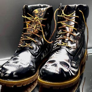 May include: Pair of black leather boots featuring skull designs and yellow laces. The boots have a glossy finish and a tan sole. The skull design is painted in white and grey. The boots are reflected on a mirrored surface.