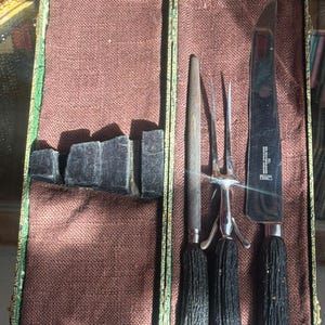 May include: An antique carving set in a green and gold case. The set includes a carving knife, a carving fork, and a sharpening steel, all with black textured handles. The case has a brown fabric lining.