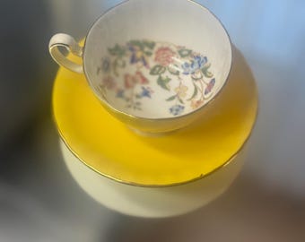 Può includere: Un set vintage di tazza e piattino da tè. La tazza presenta un motivo floreale in rosa, blu e giallo su sfondo bianco. Il piattino e la base sono giallo brillante con un bordo dorato. Il set è progettato per tè o caffè.