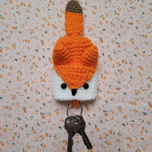 May include: A handmade crochet fox keychain. The fox has a white face, orange body, and a brown tail tip. The keychain includes a metal ring and two keys. The background is a floral pattern.