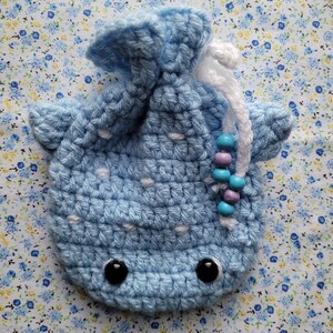 May include: A light blue crocheted whale-shaped drawstring bag with white polka dots and black eyes. The bag has a white drawstring with purple and blue beads.