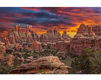 May include: A scenic landscape featuring towering red rock formations under a vibrant sunset sky. The image showcases a canyon with rugged cliffs, green vegetation, and a dramatic display of orange, yellow, and blue hues in the sky.