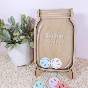 May include: A wooden mason jar-shaped display stand with a clear acrylic front panel featuring the text Brother Jar. The stand includes two small light blue and white smiley face tokens inside, with three additional tokens in light blue, pink, and peach below.