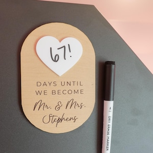 May include: A light wood oval countdown sign with a white heart-shaped acrylic insert displaying the number 67! in black marker. The sign features the engraved text DAYS UNTIL WE BECOME Mr. & Mrs. Stephens and includes a black dry erase marker.