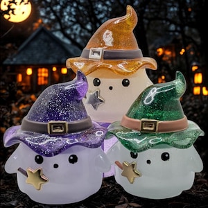 May include: Three whimsical Halloween-themed figurines. Each figure has a round body and wears a glittery witch hat in orange, purple, and green. They have black eyes, a star-shaped wand, and a silver star accent. The background features a spooky house and a full moon.
