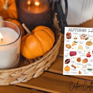 May include: A lit white candle in a glass jar, surrounded by miniature orange pumpkins and a woven basket. A printed "Autumn Bucket List" is visible, with illustrations of fall activities. The scene evokes a cozy autumn theme.