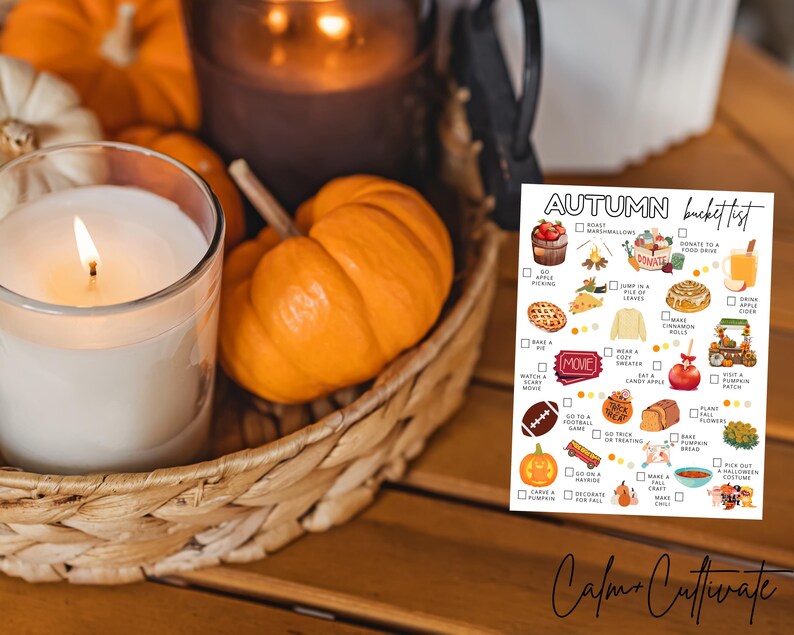 May include: A lit white candle in a glass jar, surrounded by miniature orange pumpkins and a woven basket. A printed "Autumn Bucket List" is visible, with illustrations of fall activities. The scene evokes a cozy autumn theme.