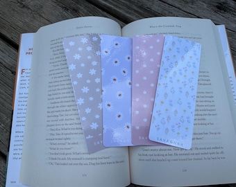 May include: Four decorative bookmarks with floral patterns and the name "SAVANNAH" printed on one. The bookmarks are inserted into an open book with text on the pages. The bookmarks have a light pink, light blue, and beige color scheme.