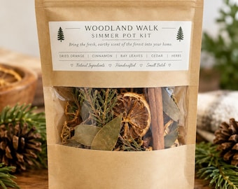 May include: A brown kraft paper pouch containing a Woodland Walk Simmer Pot Kit. The label features pine tree icons and lists ingredients including dried orange, cinnamon, bay leaves, cedar, and herbs. The clear window reveals the dried contents.