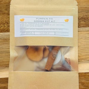 May include: A brown paper bag labeled "Pumpkin Pie Simmer Pot Kit" with a clear window showing apples, cinnamon sticks, and other ingredients. The label includes instructions and ingredient details. The bag is on a wooden surface.