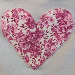 Pode incluir: Uma aplica&ccedil;&atilde;o de tecido em forma de cora&ccedil;&atilde;o com um padr&atilde;o floral em tons de rosa e branco, costurada sobre um fundo de tecido branco. O design floral apresenta rosas e folhas.