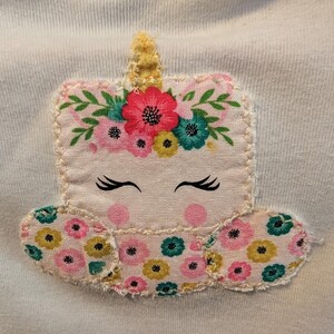 Pode incluir: Aplica&ccedil;&atilde;o de tecido de cor clara de um unic&oacute;rnio com uma coroa floral e olhos fechados. O unic&oacute;rnio tem um chifre amarelo e &eacute; decorado com flores rosa, amarelas e turquesas. A aplica&ccedil;&atilde;o &eacute; costurada em um tecido azul claro.