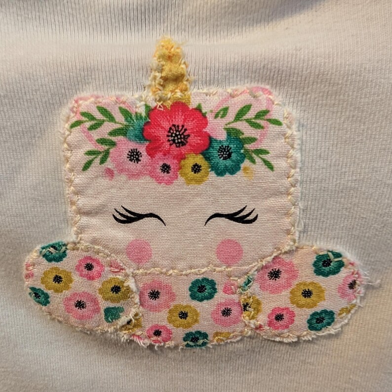 Pode incluir: Aplica&ccedil;&atilde;o de tecido de cor clara de um unic&oacute;rnio com uma coroa floral e olhos fechados. O unic&oacute;rnio tem um chifre amarelo e &eacute; decorado com flores rosa, amarelas e turquesas. A aplica&ccedil;&atilde;o &eacute; costurada em um tecido azul claro.