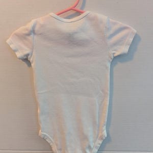 Pode incluir: Um body branco para beb&eacute; com mangas curtas est&aacute; pendurado num cabide de pl&aacute;stico rosa. O body tem um decote redondo e &eacute; feito de um tecido macio e de cor clara. A pe&ccedil;a de roupa &eacute; projetada para beb&eacute;s.