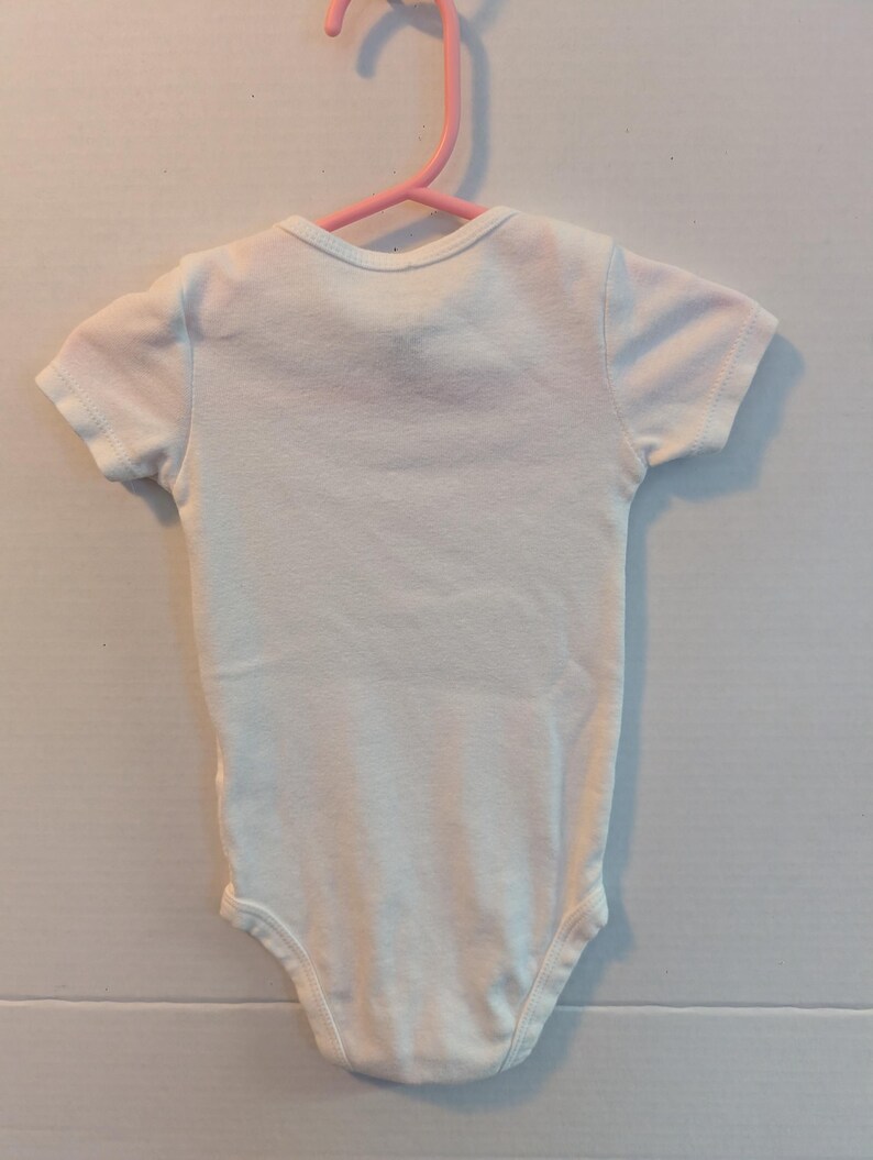 Pode incluir: Um body branco para beb&eacute; com mangas curtas est&aacute; pendurado num cabide de pl&aacute;stico rosa. O body tem um decote redondo e &eacute; feito de um tecido macio e de cor clara. A pe&ccedil;a de roupa &eacute; projetada para beb&eacute;s.