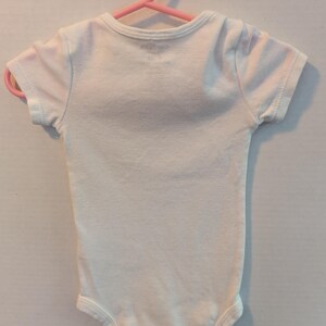 Pode incluir: Um body branco para beb&eacute; com mangas curtas, pendurado num cabide de pl&aacute;stico rosa. A pe&ccedil;a de roupa &eacute; feita de um tecido macio. O body &eacute; projetado para beb&eacute;s e crian&ccedil;as pequenas.