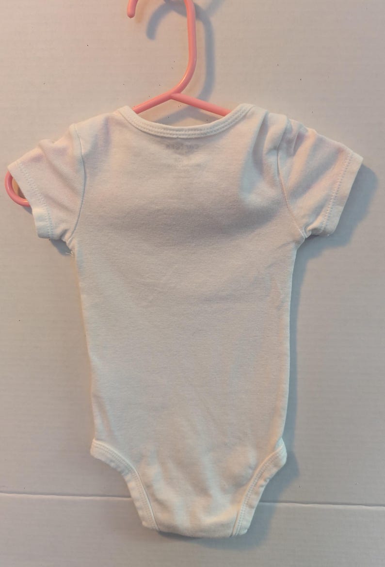 Pode incluir: Um body branco para beb&eacute; com mangas curtas, pendurado num cabide de pl&aacute;stico rosa. A pe&ccedil;a de roupa &eacute; feita de um tecido macio. O body &eacute; projetado para beb&eacute;s e crian&ccedil;as pequenas.