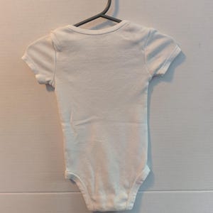 Pode incluir: Um body branco para beb&eacute; com mangas curtas, pendurado num cabide cinzento. O body &eacute; feito de tecido macio e tem um design simples. Esta pe&ccedil;a de roupa &eacute; para beb&eacute;s e crian&ccedil;as pequenas.