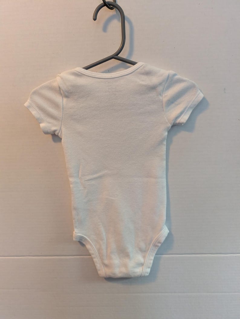 Pode incluir: Um body branco para beb&eacute; com mangas curtas, pendurado num cabide cinzento. O body &eacute; feito de tecido macio e tem um design simples. Esta pe&ccedil;a de roupa &eacute; para beb&eacute;s e crian&ccedil;as pequenas.