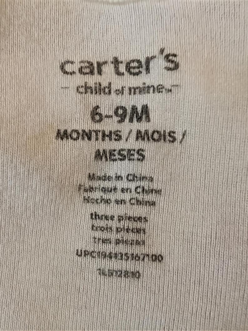 Pode incluir: Close-up de uma etiqueta de tecido branco com o texto "carter's - child of mine" e informa&ccedil;&otilde;es de tamanho para beb&ecirc;s de 6 a 9 meses. A etiqueta tamb&eacute;m inclui o texto "Made in China" e "three pieces".