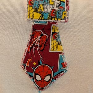 Pode incluir: Camiseta branca com um patch costurado colorido com as palavras "WALL CRAWLER" em vermelho, amarelo e azul. Abaixo do texto, h&aacute; um patch em forma de diamante com um design do Homem-Aranha, incluindo sua m&aacute;scara e uma m&atilde;o lan&ccedil;ando uma teia.