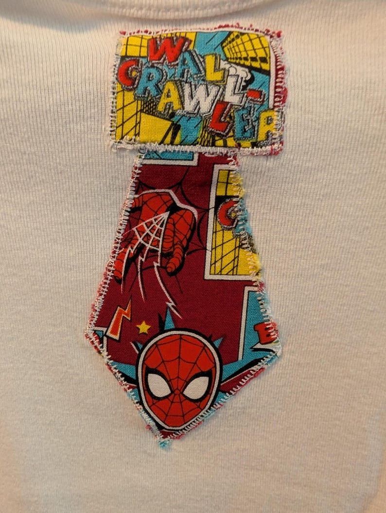 Pode incluir: Camiseta branca com um patch costurado colorido com as palavras "WALL CRAWLER" em vermelho, amarelo e azul. Abaixo do texto, h&aacute; um patch em forma de diamante com um design do Homem-Aranha, incluindo sua m&aacute;scara e uma m&atilde;o lan&ccedil;ando uma teia.