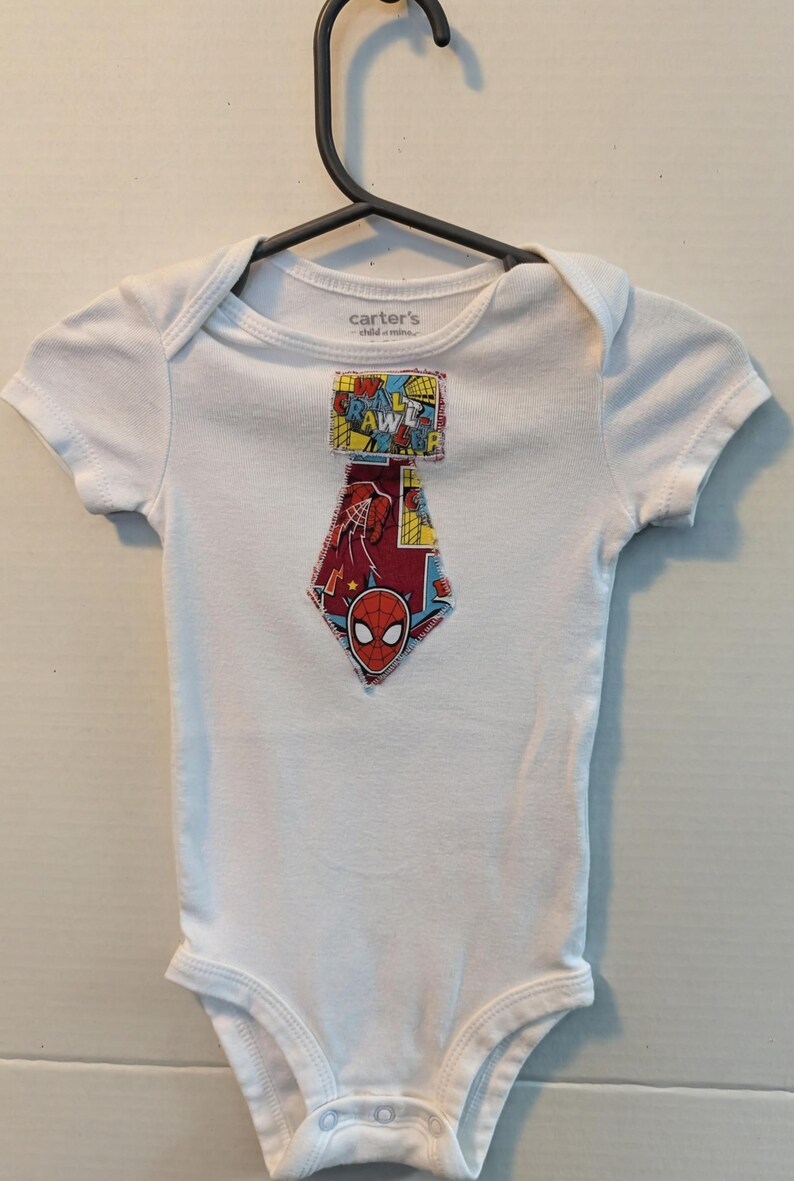 Pode incluir: Body de beb&eacute; branco com um gr&aacute;fico do Homem-Aranha. O gr&aacute;fico apresenta a m&aacute;scara do Homem-Aranha e as palavras "Crawl" e "Awesomer" em estilo de banda desenhada. O body est&aacute; pendurado num cabide preto.