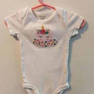 Pode incluir: Body de beb&ecirc; branco com um design de unic&oacute;rnio. O unic&oacute;rnio tem um rosto rosa, um chifre dourado e uma crina floral. O body est&aacute; em um cabide rosa. A marca "Carter's" &eacute; vis&iacute;vel na etiqueta.